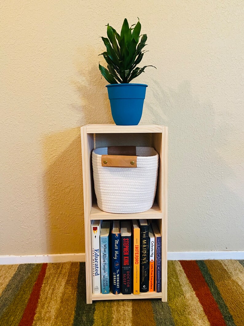 Op de afbeelding: Een licht houten plank met twee planken. De bovenste plank heeft een witte touwenmand met een bruin leren handvat. Een blauwe pot met een groene plant staat bovenop de plank. De onderste plank heeft een rij boeken met titels waaronder "Educated", "What Alice Forgot", "The Midnight Library", "Matt Haig", "The Maidens", "Inferno", "Doctor Sleep", en "Glass Castle".