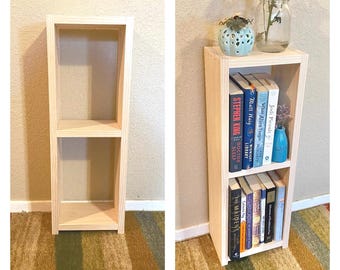May include: A tall, rectangular, light-colored wooden bookshelf featuring two open storage compartments. The unit is constructed from unfinished wood planks with visible grain, providing a simple, functional structure for organizing books and decorative items.