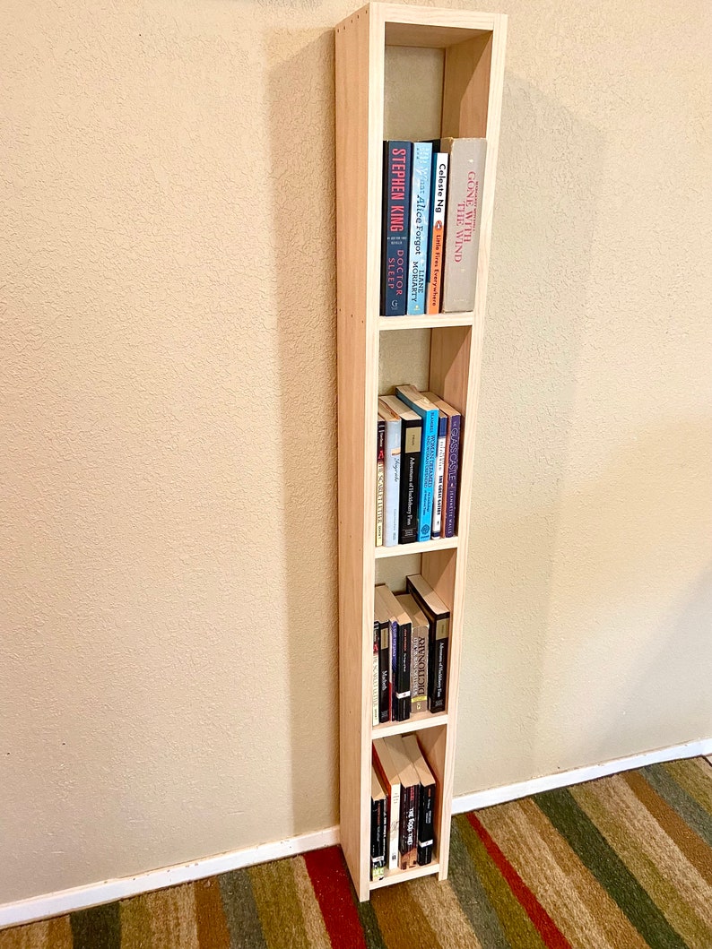 May include: A light wood bookshelf with four shelves, filled with books. The bookshelf is standing against a light beige wall. The books on the top shelf include "Gone With The Wind", "Alice Forgot", "The Time Traveler's Wife", "Doctor Sleep", and "Monday's Child".