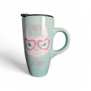 May include: A light teal travel mug with a black lid and handle. The mug features a white cat face wearing pink heart-shaped glasses with blue eyes. The words "CRAZY CAT LADY" are printed in pink.