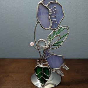 May include: A stained glass fairy figurine with a lavender flower, green leaf, and a pink bead. The fairy has lavender wings and a silver metal frame. The figurine is on a silver metal base and sits on a brown surface.