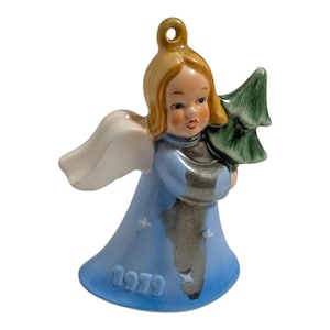 May include: A ceramic angel Christmas ornament. The angel has blonde hair, a blue dress with the year "1979" printed on it, and white wings. The angel is holding a green Christmas tree. The ornament has a gold loop for hanging.
