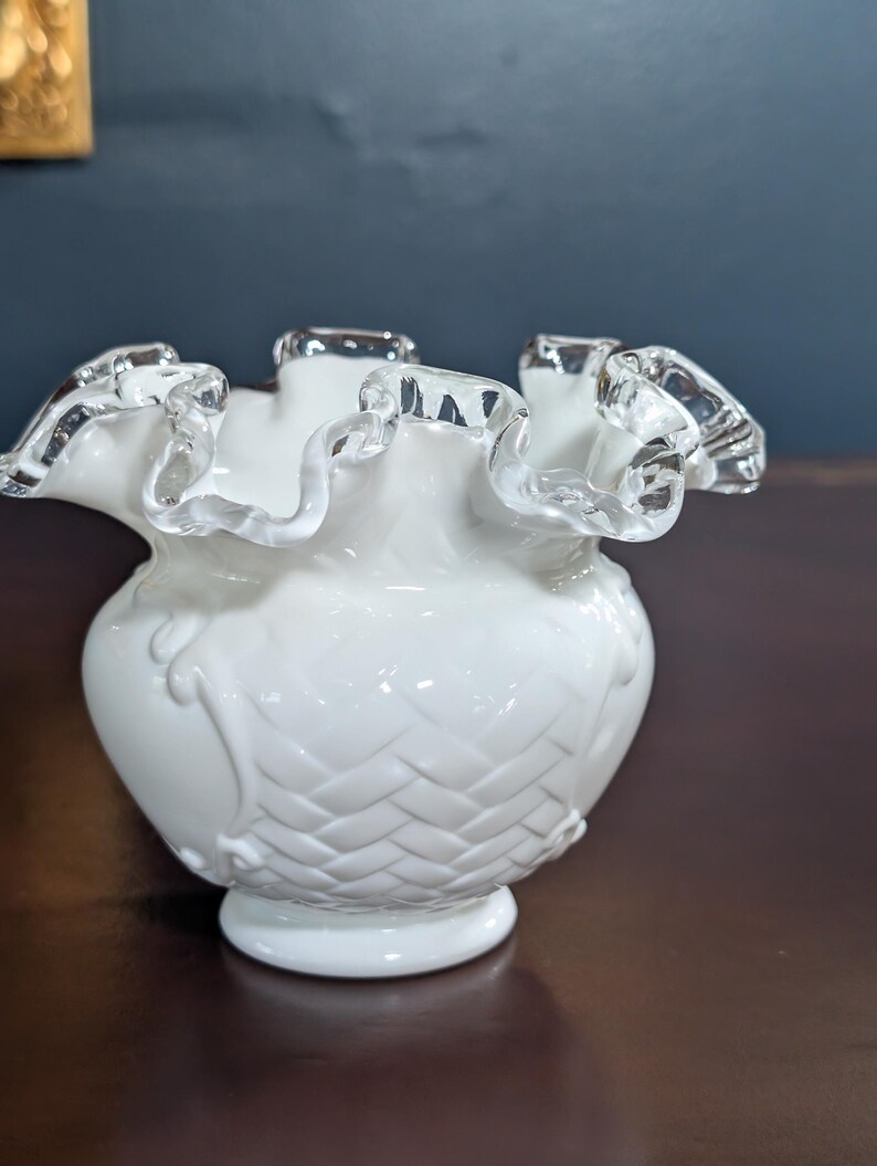 May include: A white milk glass vase with a ruffled top and a woven pattern. The vase has a silver trim around the top.