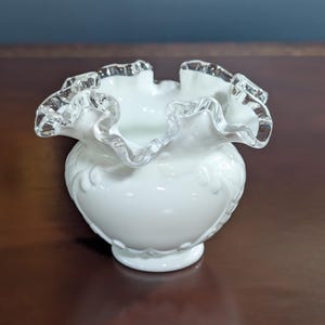 May include: A white milk glass vase with a ruffled, silver-trimmed rim. The vase has a delicate, embossed design.