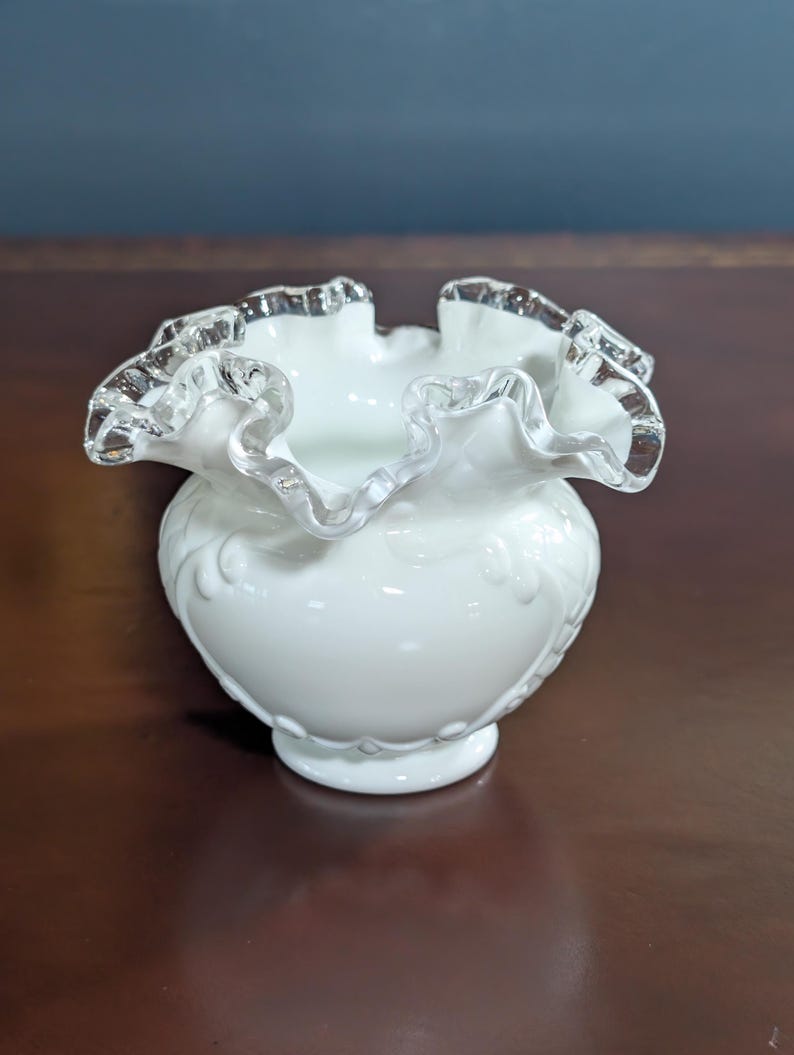 May include: A white milk glass vase with a ruffled, silver-trimmed rim. The vase has a delicate, embossed design.