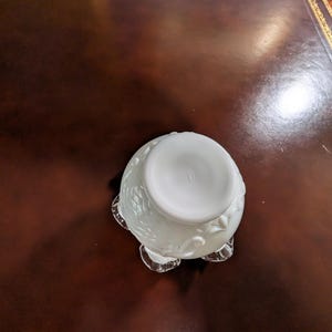 May include: White ceramic bowl with a floral design and a scalloped edge. The bowl is sitting on a brown leather surface.