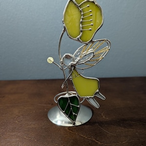 May include: A stained glass fairy sculpture with a yellow flower and green leaf. The fairy has yellow wings and a silver body. The sculpture is on a silver base and has a small yellow bead.