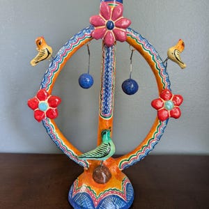 May include: A colorful, hand-painted decorative sculpture featuring a circular design with orange, blue, and red accents. The sculpture includes two small yellow birds, two red flowers, and two blue hanging spheres. A green bird is perched on the base.