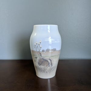 May include: A white ceramic vase with a landscape design. The vase features a pastoral scene with a large rock, grass, and trees. The vase is approximately 6 inches tall.
