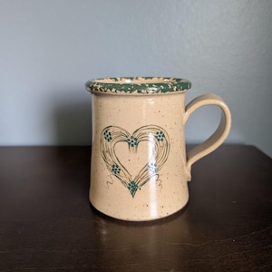 May include: A beige ceramic mug with a speckled finish and a dark green rim. The front of the mug features a hand-painted dark green heart design composed of swirling lines and small dots. The mug has a single curved handle on the right side.