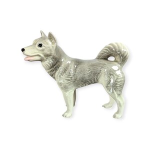 May include: A ceramic figurine of a dog in a standing pose. The dog has gray and white fur, with a curled tail and a pink tongue. The figurine is on a white background.