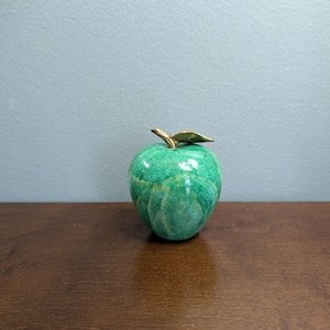 May include: A decorative apple figurine featuring a glossy, mottled green ceramic body with subtle variations in tone. The apple is topped with a metallic gold-colored stem and a single leaf, providing a distinct contrast to the textured green surface.
