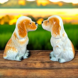 May include: Two white ceramic figurines of cocker spaniels facing each other. The dogs are brown and white and have floppy ears. The figurines are on a wooden surface.