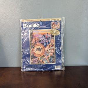 May include: A Bucilla needlepoint kit titled "Indian and Buffalo." The kit is in a clear plastic package and features a colorful design with Native American imagery, including a buffalo and a warrior. The package measures 13 x 14 inches.