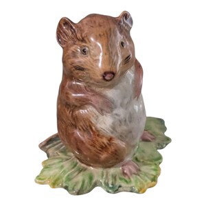 May include: A brown and white ceramic figurine of a mouse sitting on a green leaf. The mouse has a brown body, white belly, and pink nose.