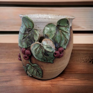 May include: A handcrafted ceramic vase with a textured, speckled beige rim and a brown body. The vase is adorned with raised green leaves and clusters of maroon berries, creating a nature-inspired design. The vase is suitable for home decor.