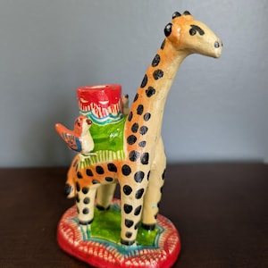 May include: A hand-painted giraffe figurine with a red, green, and blue saddle. The giraffe has black spots and a long neck. The base is red with a green and blue design. A small bird is perched on the giraffe's back.