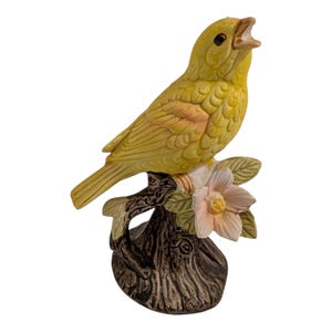 May include: A yellow ceramic bird figurine perched on a brown tree branch with pink and white flowers. The bird has an open beak and is facing to the right. The base of the figurine is brown.