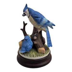 May include: A ceramic figurine featuring two blue jays perched on a brown tree branch. The larger bird has a crest and is detailed with blue and white feathers. The base is a dark brown, round shape with a touch of green grass.