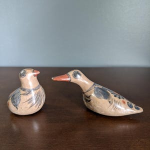 May include: Two ceramic bird figurines. The birds are tan with black and blue painted details. One bird is upright, and the other is in a resting position. Both have red beaks and silver eyes. The figurines are on a dark brown surface.