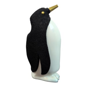 May include: A white ceramic penguin figurine with a black felt body and a gold beak. The penguin is standing upright with its wings folded.