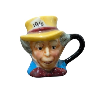 May include: A ceramic mug shaped like the Mad Hatter from Alice in Wonderland. The mug features a yellow hat with the text "10/6", a red bow tie, and a black handle. The face is detailed with painted features.