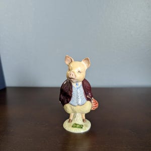 May include: A ceramic figurine of a pig standing upright, dressed in a dark maroon jacket, a light blue vest, and cream-colored trousers. The pig holds a small red patterned bag in one hand and stands on a circular base with a green Beswick England stamp.