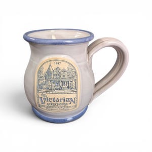 May include: A light gray ceramic mug with a blue rim and base, featuring a central cream-colored label with a blue illustration of a house, the text 1887, The Victorian Guest House, and Nappanee, Indiana, with a single handle on the right side.