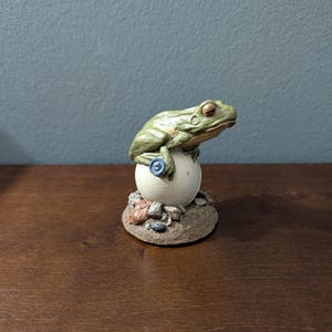 May include: A green frog figurine perched atop a smooth white sphere. The frog has textured skin and is holding a small blue circular object. The base is a textured brown mound featuring small rocks and a leaf, with the text "1995" visible on the front.