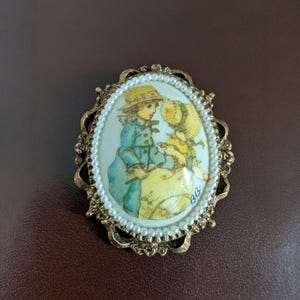 May include: Vintage oval brooch featuring a painted illustration of two children in pastel clothing. The image is framed with faux pearls and a decorative gold-tone metal border. The brooch is a classic accessory.