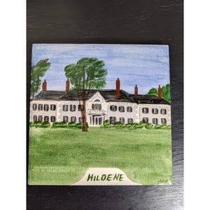 May include: A hand-painted ceramic tile depicting a white house with a green lawn and blue sky. The house has a black roof and white trim. There are two trees in the front yard. The tile is signed "Hildene" in the bottom right corner.