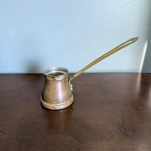 May include: A copper Turkish coffee pot with a wide, rounded base and a narrow neck. The pot features a long, flat, brass-colored metal handle attached to the side with two rivets. The interior of the pot is silver-toned metal, and the exterior has a warm, metallic copper finish with visible signs of age and use.