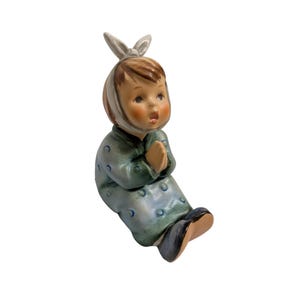May include: A vintage porcelain figurine of a seated child with hands clasped in prayer. The child wears a green dress with blue polka dots and a white head covering with bunny ears. The figurine has a serene expression.