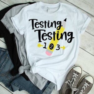 Puede incluir: Camiseta blanca con texto negro que dice "Testing Testing 1 2 3" y un lápiz amarillo con una goma rosa.