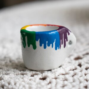 May include: A small, white ceramic cup with a handle. The rim is painted with a rainbow of colors, and the paint drips down the sides. The cup is sitting on a white, textured surface.