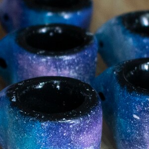May include: A collection of small, handmade ceramic mugs. Each mug features a galaxy-inspired design with shades of blue, purple, and white. The interior of each mug is black. The mugs are arranged on a wooden surface.