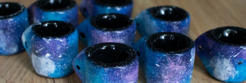 May include: A collection of small, handmade ceramic mugs. Each mug features a galaxy-inspired design with shades of blue, purple, and white. The interior of each mug is black. The mugs are arranged on a wooden surface.