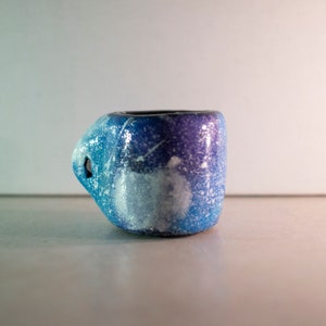 May include: A small, handmade ceramic mug with a unique handle design. The mug features a gradient of blue and purple hues, speckled with white, and a dark interior. The handle has a hole. The mug is likely used for drinking beverages.