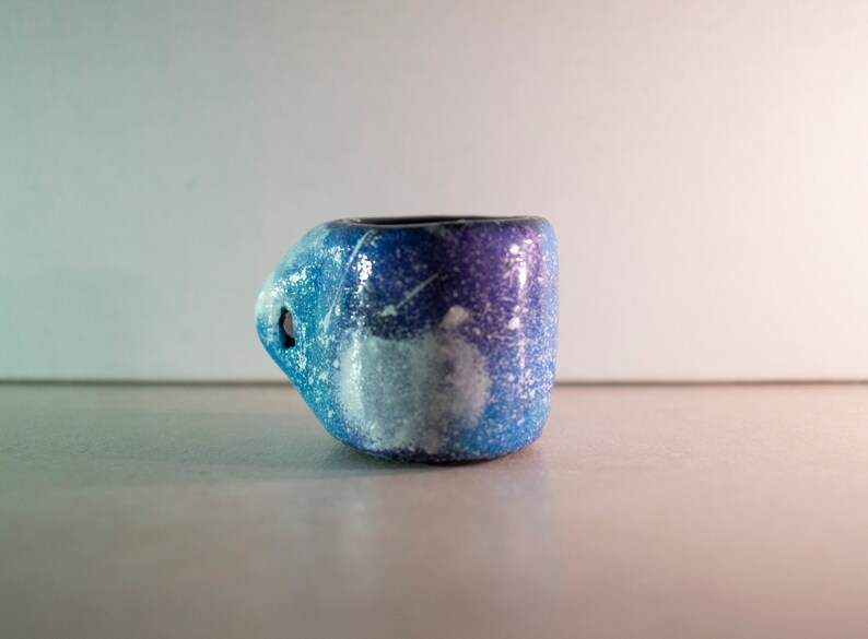 May include: A small, handmade ceramic mug with a unique handle design. The mug features a gradient of blue and purple hues, speckled with white, and a dark interior. The handle has a hole. The mug is likely used for drinking beverages.