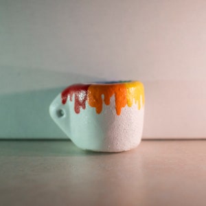 May include: A small, white ceramic mug with a handle. The top rim of the mug is painted with a gradient of colors, transitioning from red to orange and yellow, with drips cascading down the sides. The mug has a textured surface.