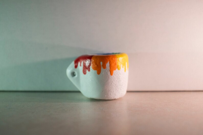 May include: A small, white ceramic mug with a handle. The top rim of the mug is painted with a gradient of colors, transitioning from red to orange and yellow, with drips cascading down the sides. The mug has a textured surface.