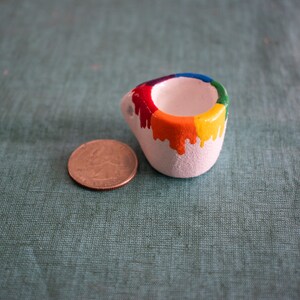 May include: A small, white ceramic container with a rainbow-colored paint drip design. The colors include red, orange, yellow, green, and blue. A U.S. quarter coin is next to the container for size comparison. The container has a rounded shape.