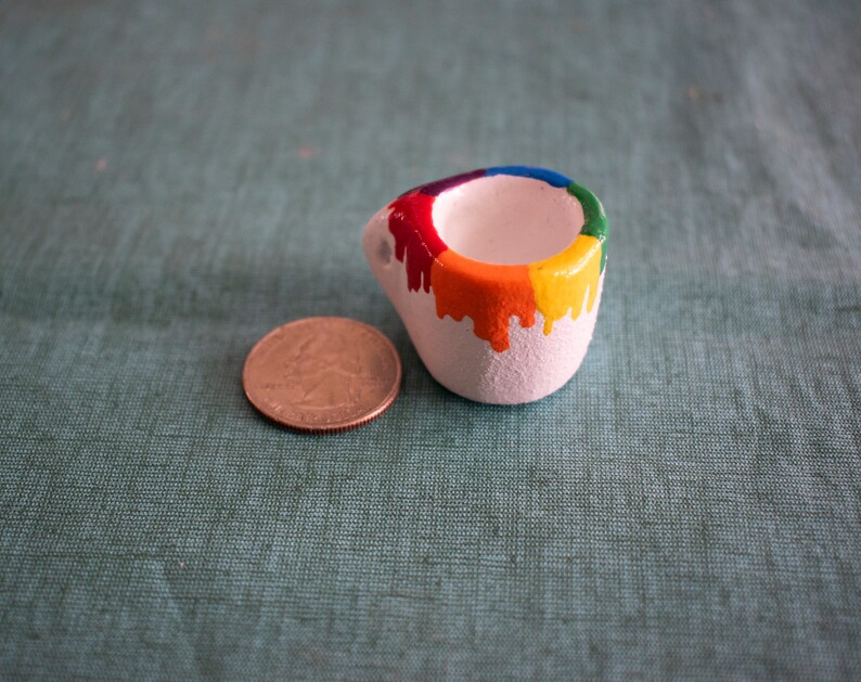 May include: A small, white ceramic container with a rainbow-colored paint drip design. The colors include red, orange, yellow, green, and blue. A U.S. quarter coin is next to the container for size comparison. The container has a rounded shape.