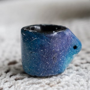 May include: A small, cylindrical ceramic ring holder with a dark interior and a gradient of blue and purple hues. The ring holder has a small, rounded protrusion with a hole on one side. The surface has a speckled, textured appearance.