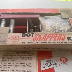 May include: Vintage Dritz Dot Snappers Kit in a cardboard box. The kit includes tools and fasteners for attaching snaps to clothing and other fabrics. The box has the text "Easy to attach at home".