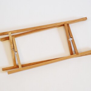 May include: A wooden frame made of four pieces of wood that are connected at the corners. The frame is folded up and is lying flat on a white surface.