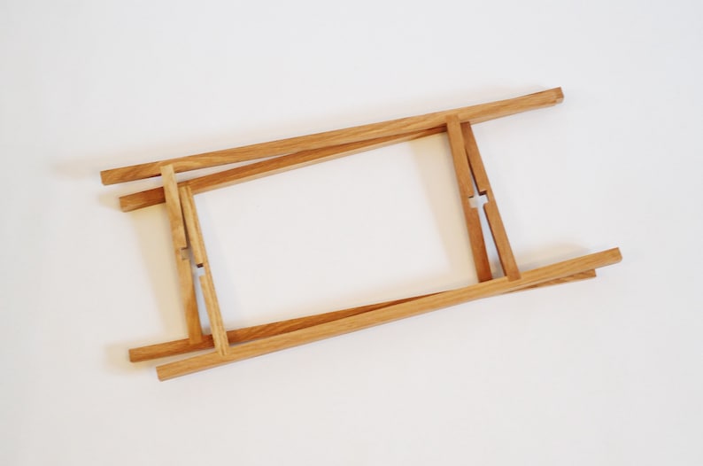 May include: A wooden frame made of four pieces of wood that are connected at the corners. The frame is folded up and is lying flat on a white surface.