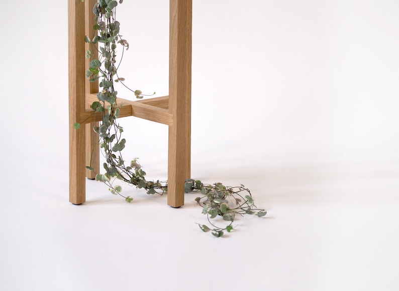 May include: A wooden plant stand with a natural finish. A trailing plant with green leaves hangs over the side of the stand.