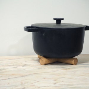 May include: A black cast iron pot with a lid, sitting on a wooden trivet. The pot has two side handles and a single handle on the lid. The trivet is made of light-coloured wood and has an X-shaped design.
