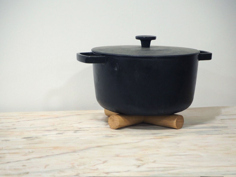 May include: A black cast iron pot with a lid, sitting on a wooden trivet. The pot has two side handles and a single handle on the lid. The trivet is made of light-coloured wood and has an X-shaped design.